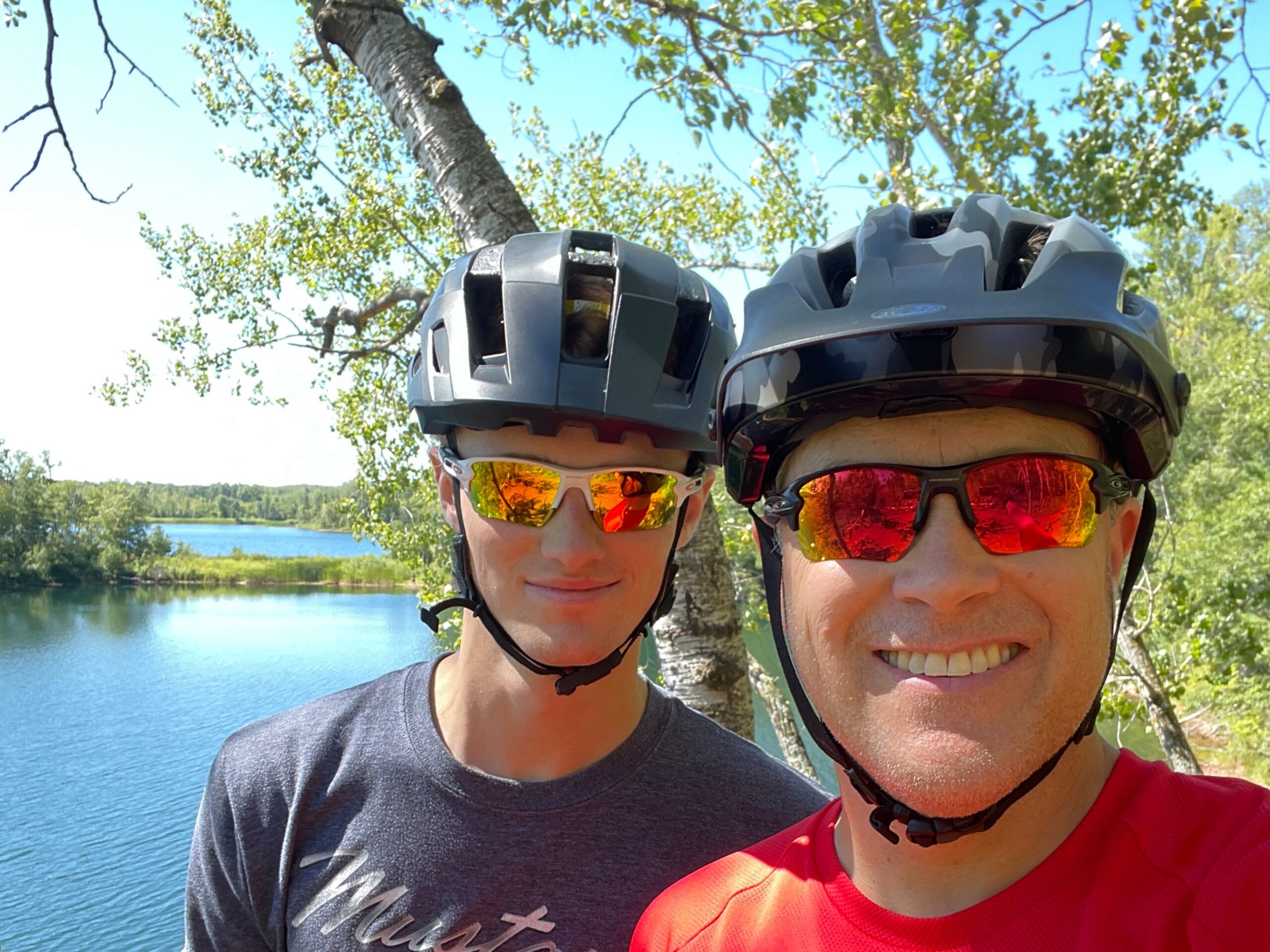 Matt Bischel and his son smiling in front of a lake and birch trees with bike helmets on their heads