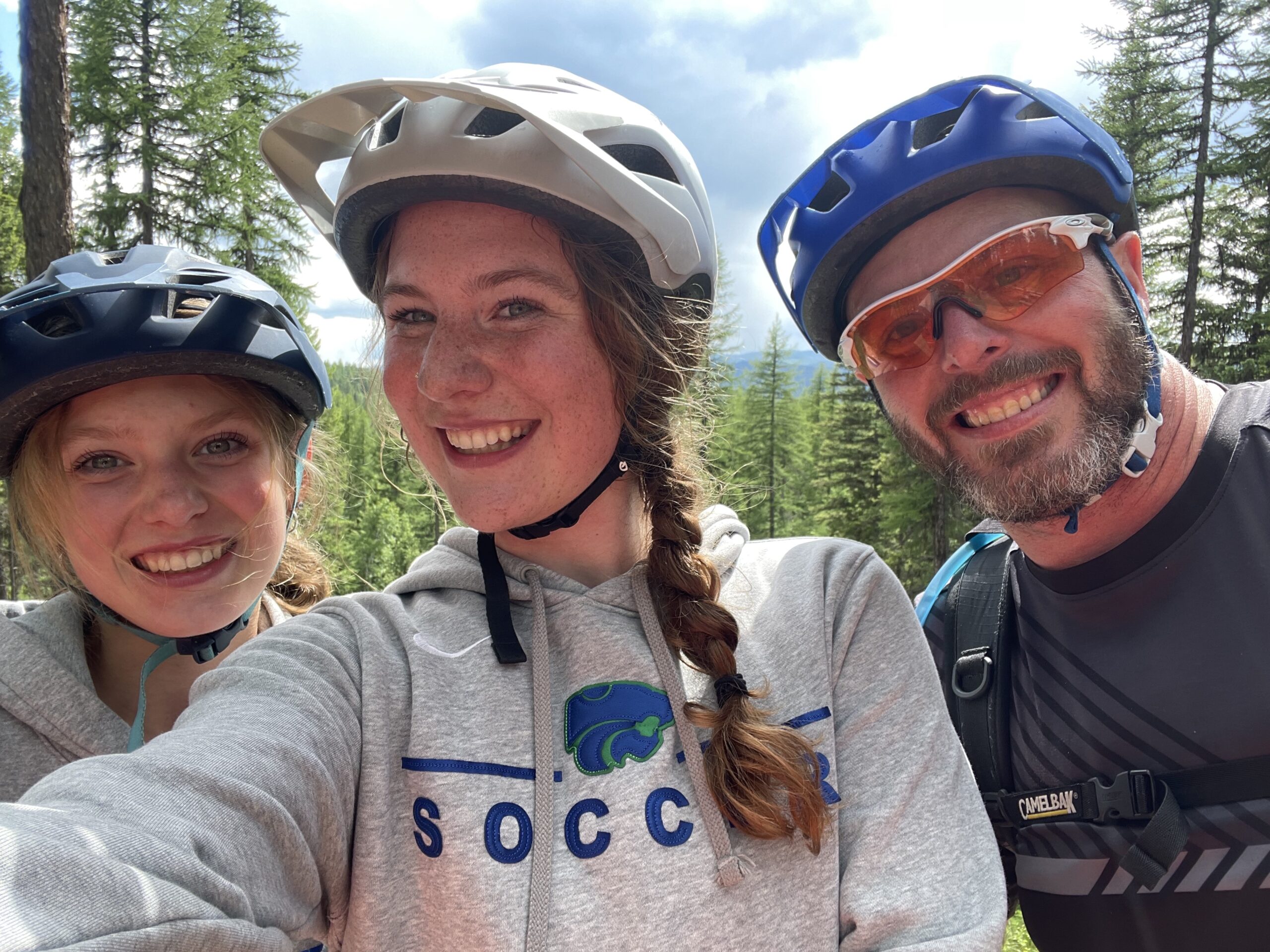 Reid Bartels stands smiling with his two daughters, all wearing bike helmets.